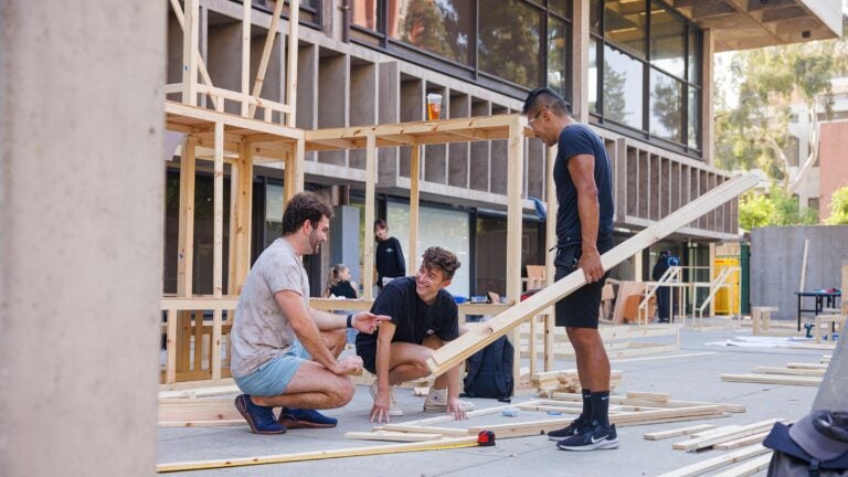 Three students talk together as they build a wooden structure and other partially built structures stand in the back at the USC School of Architecture.